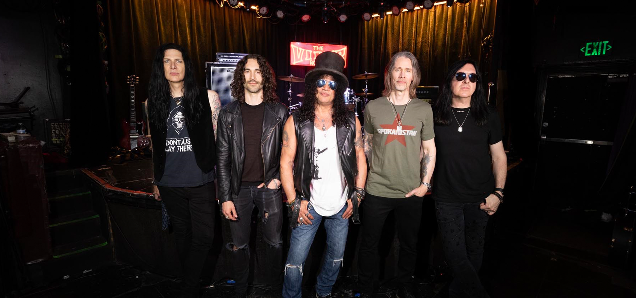 Slash featuring Myles Kennedy and the Conspirators posing together on a dimly lit music venue stage.