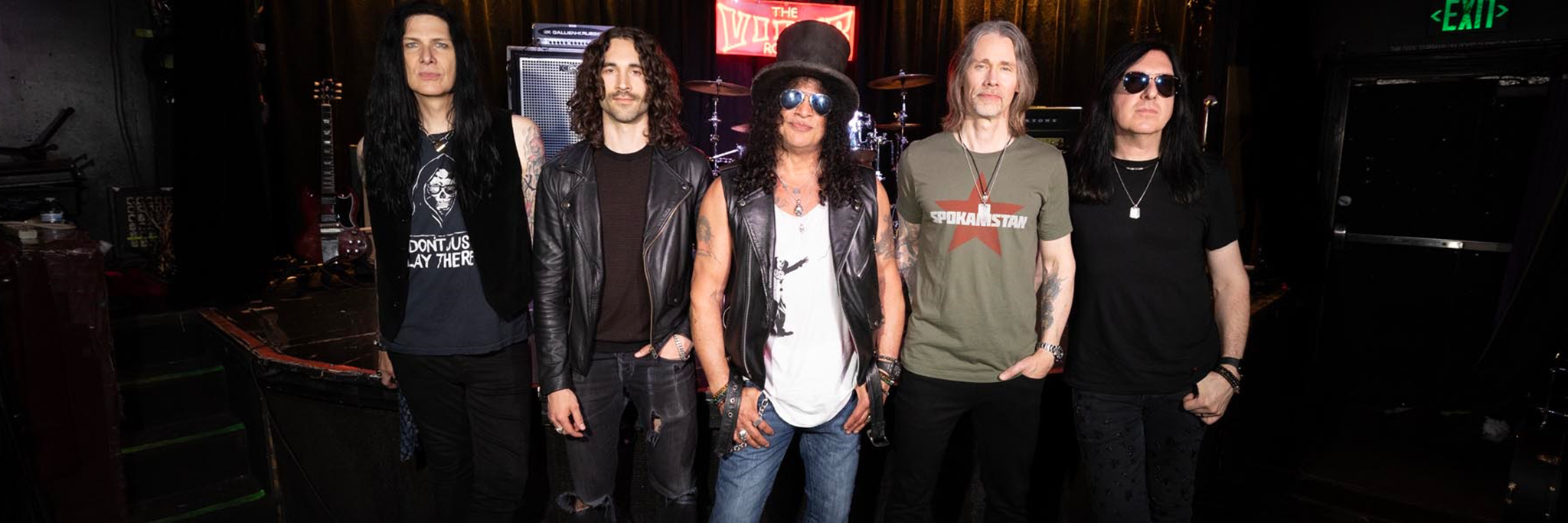 Slash featuring Myles Kennedy and the Conspirators posing together on a dimly lit music venue stage.