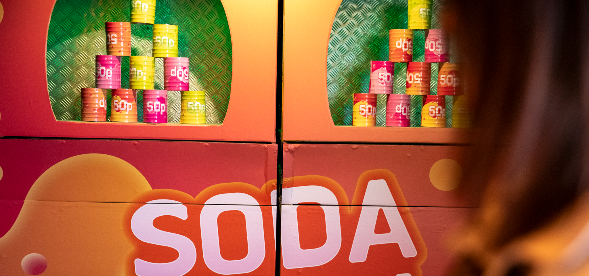 Staplar av färgglada burkar märkta "50p" visas i ett stånd med ordet "SODA" tryckt under; en person syns delvis i förgrunden.