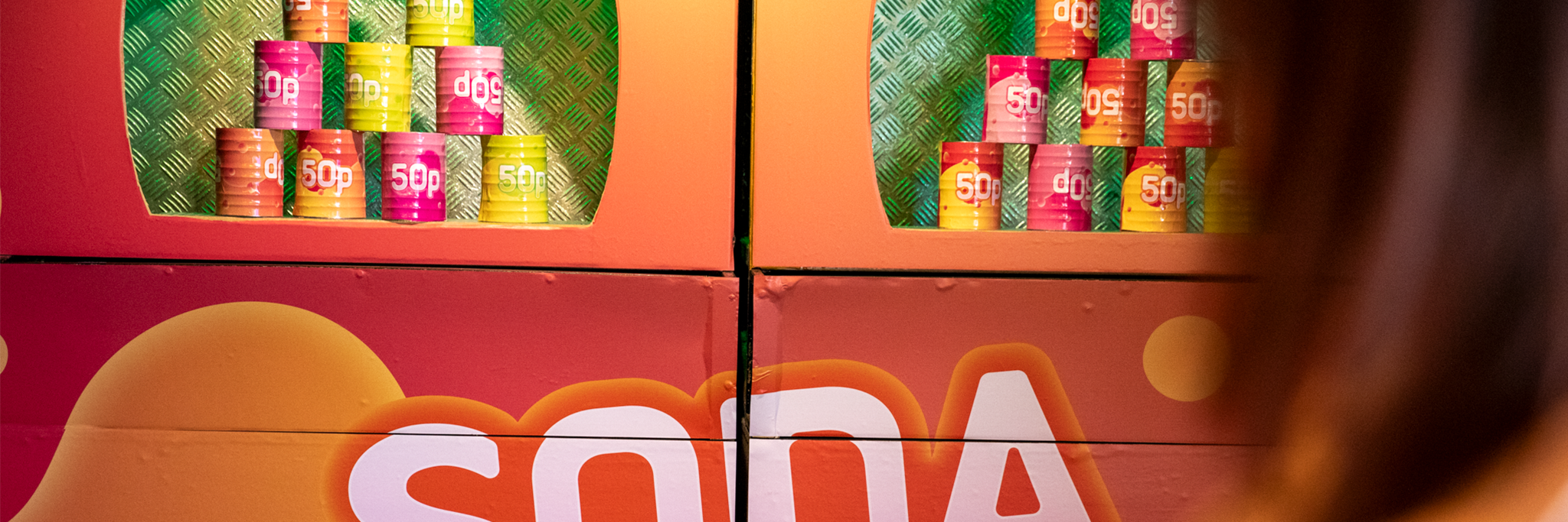 Staplar av färgglada burkar märkta "50p" visas i ett stånd med ordet "SODA" tryckt under; en person syns delvis i förgrunden.