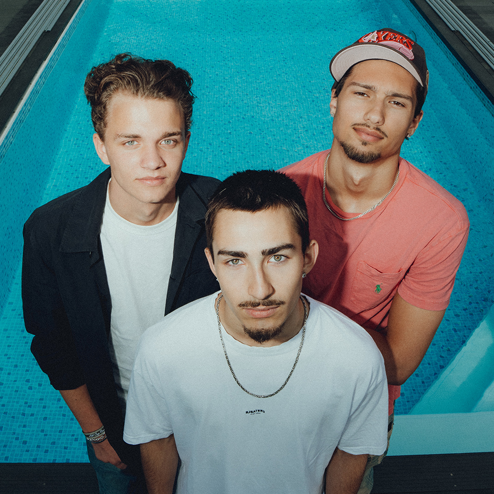 Three young men stand closely together at the edge of a swimming pool, looking up at the camera.