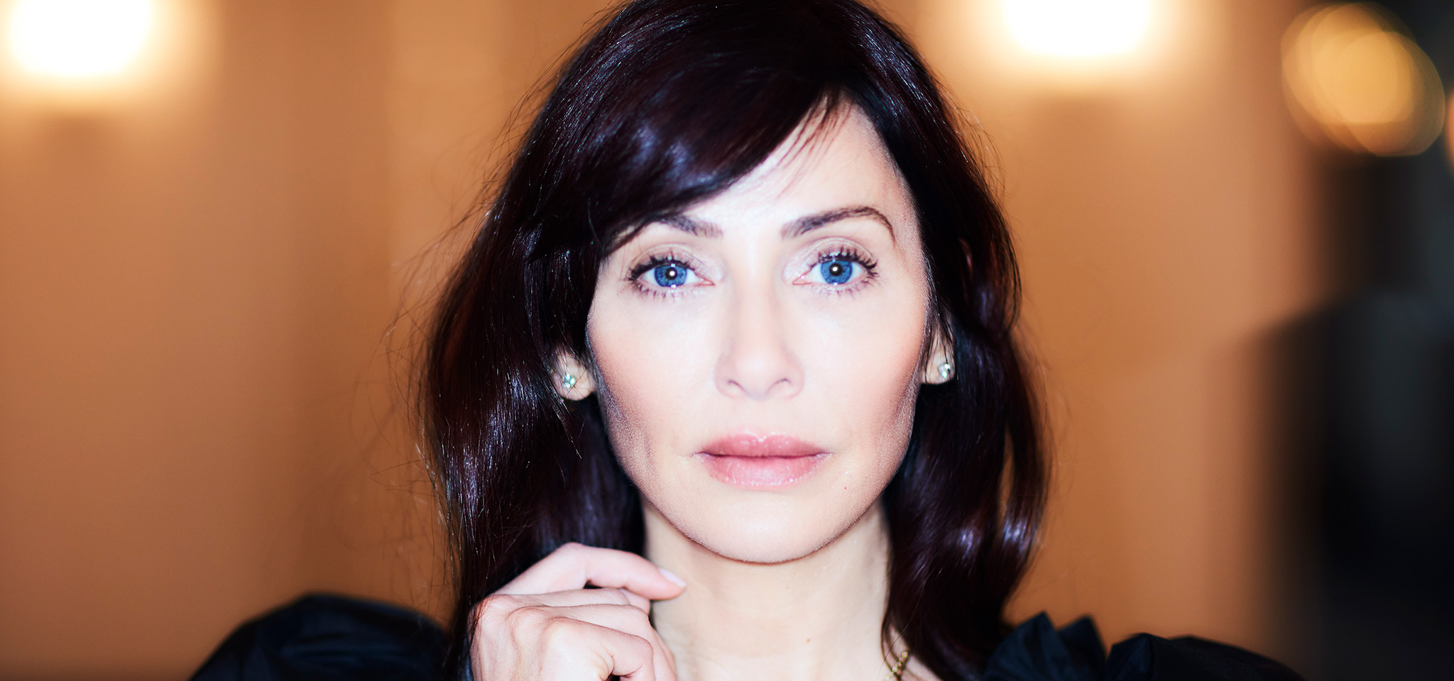 En kvinna med långt mörkt hår och blå ögon tittar rakt in i kameran, klädd i en svart topp, med en neutral bakgrund och mjuk belysning.