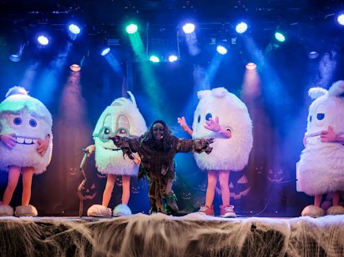 Performer in a ragged costume poses on stage in front of three people wearing large, white, furry character costumes, with stage lights and fog effects in the background.