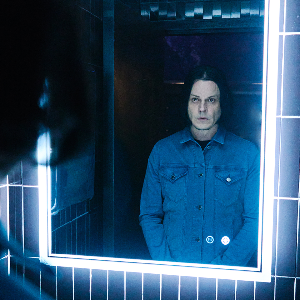 A person with shoulder-length dark hair and a blue denim jacket stands in front of a lit bathroom mirror, looking at their reflection.
