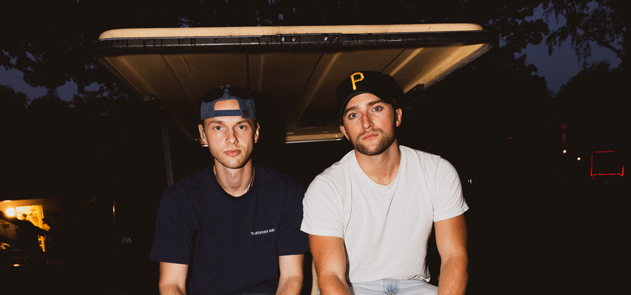 Två män sitter sida vid sida på kanten av en golfbil på kvällen. Den ene har en bakvänd keps och mörk skjorta, den andre har en svart keps med ett "P" och en vit t-shirt.