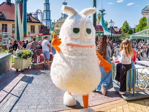En person i en stor, fluffig vit maskotdräkt med orange armar går genom en fullsatt nöjespark en solig dag.