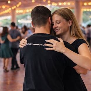 Ett ungt par dansar tätt tillsammans på ett inomhusevenemang, med andra par synliga i bakgrunden under ljusslingor.
