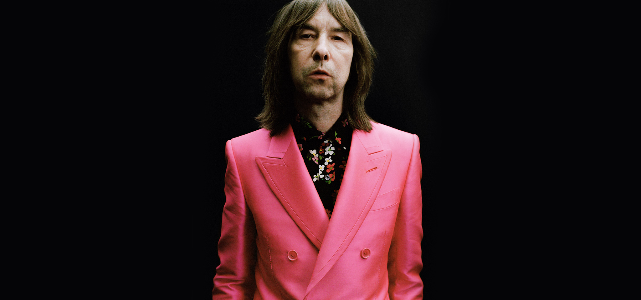 En person med långt brunt hår bär en ljusrosa kostymjacka och en svartblommig skjorta och står mot en svart bakgrund.