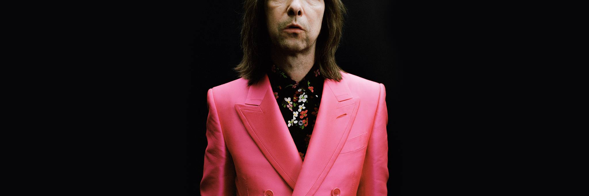 En person med långt brunt hår bär en ljusrosa kostymjacka och en svartblommig skjorta och står mot en svart bakgrund.