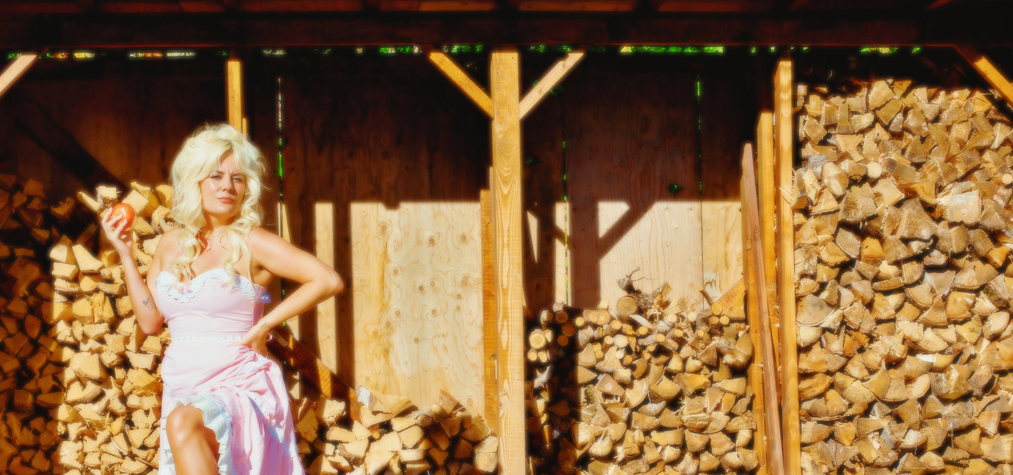 En kvinna i ljusrosa klänning står framför en staplad vedtrave och håller i ett äpple under ett träskydd en solig dag.