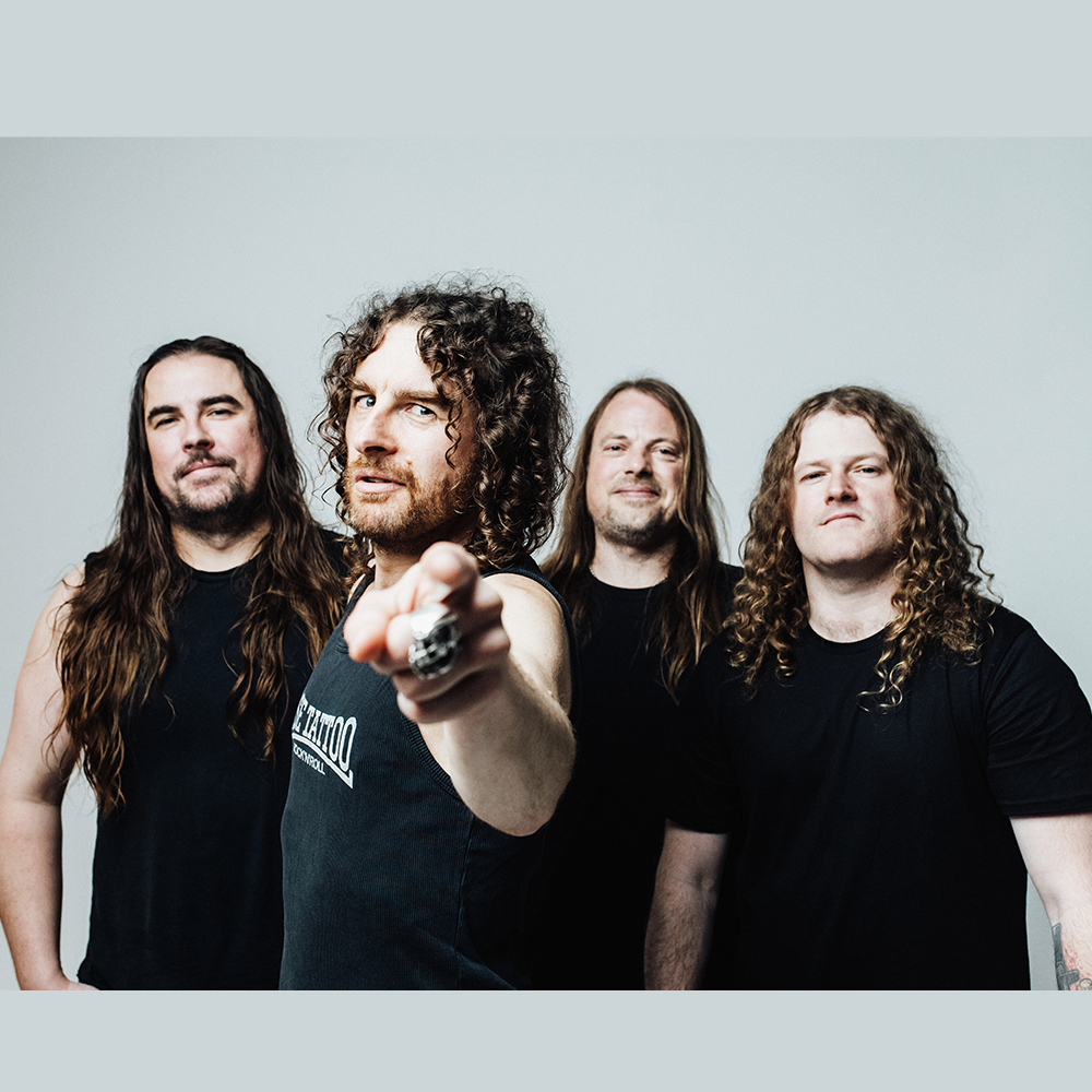 Fyra män med långt hår, klädda i svarta skjortor, står tillsammans framför en vanlig ljus bakgrund. Mannen längst fram pekar mot kameran.