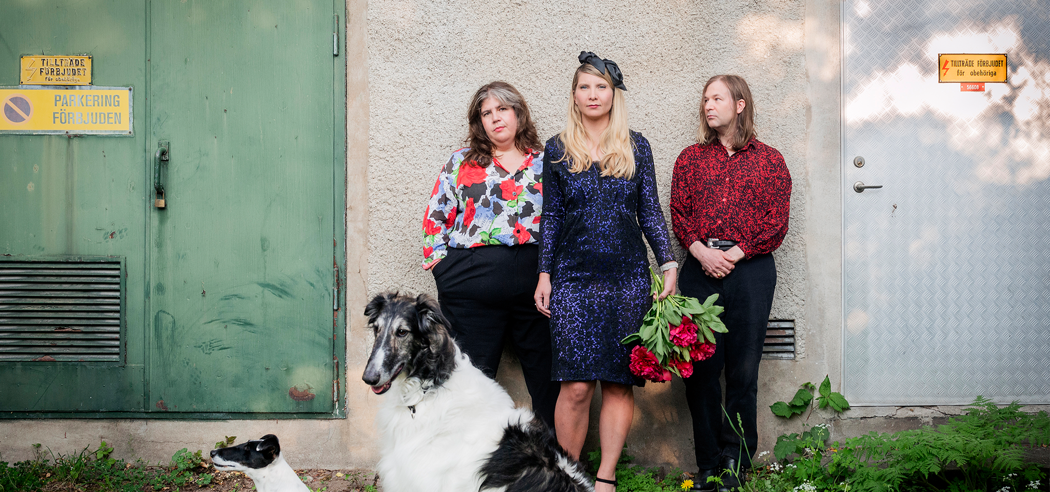 Tre personer står mot en vägg nära en dörr, med två hundar i förgrunden. En person håller i en bukett med röda blommor. Runt omkring dem syns skyltar och grönska.