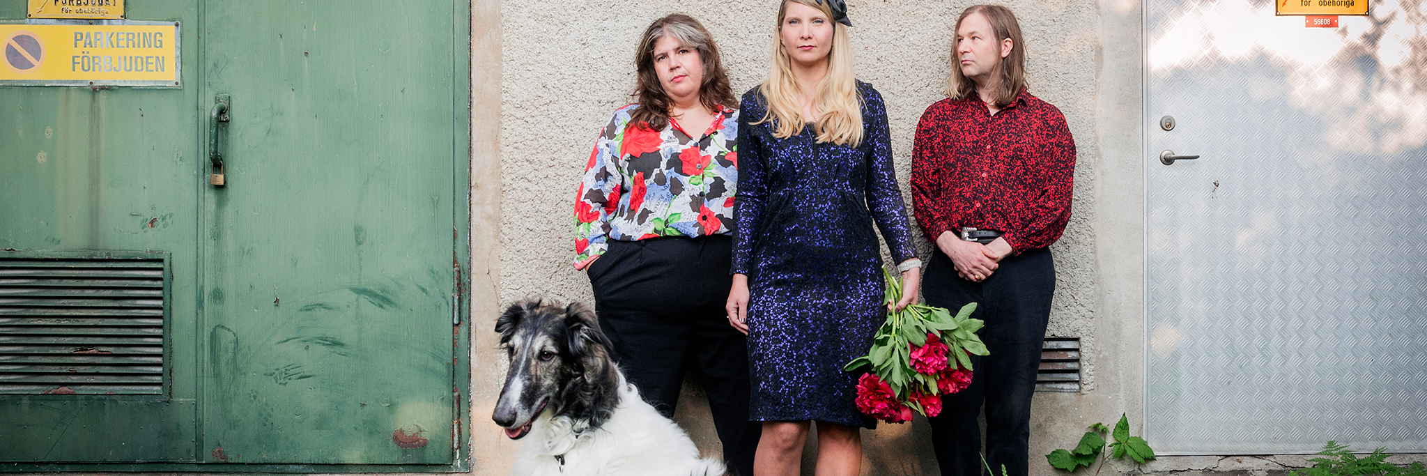 Tre personer står mot en vägg nära en dörr, med två hundar i förgrunden. En person håller i en bukett med röda blommor. Runt omkring dem syns skyltar och grönska.