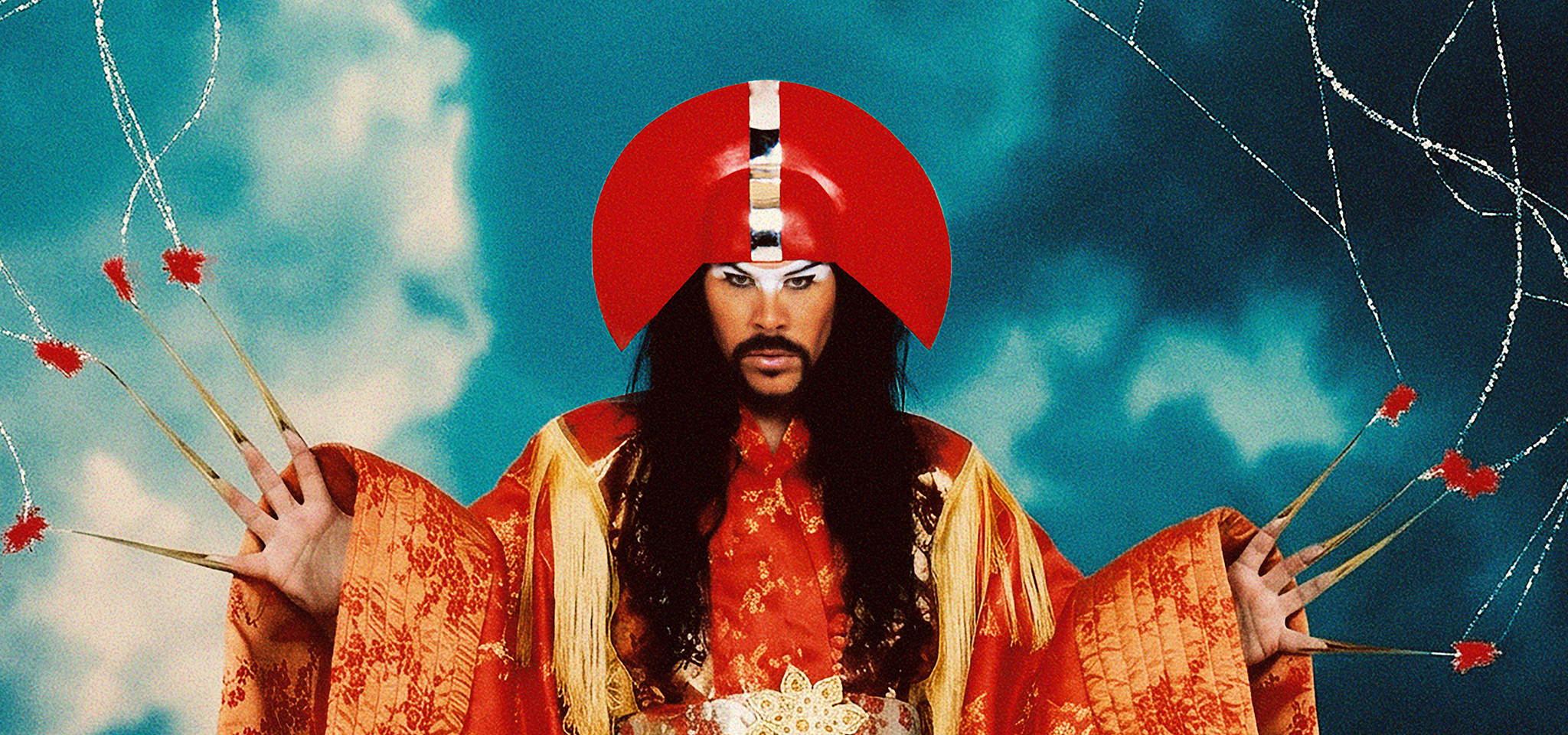 En person iförd en utsmyckad dräkt i rött och guld med långa spetsiga naglar och en stor röd huvudbonad poserar med händerna höjda mot en bakgrund av molnig himmel och taggtråd.