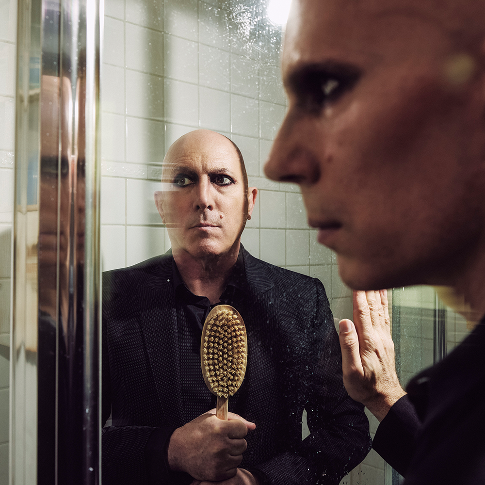 A person with heavy eyeliner looks into a mirror, reflecting another person holding a wooden brush, both in a tiled bathroom.