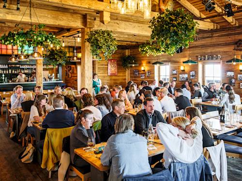 A busy rustic restaurant with wooden decor and hanging plants, where many people are seated at tables eating, drinking, and talking. A server stands near the back, ready to take orders.