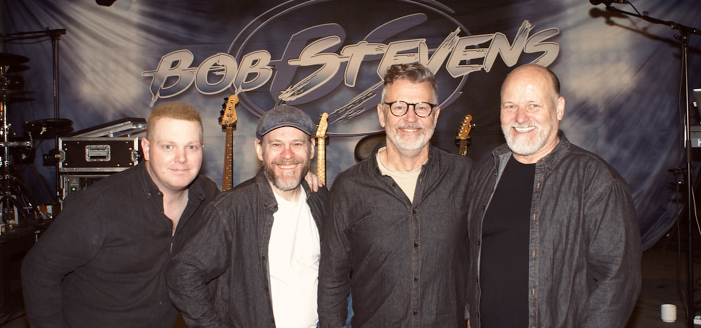 Fyra män står framför en kuliss med texten "Bob Stevens" och bakom dem syns musikinstrument och utrustning.