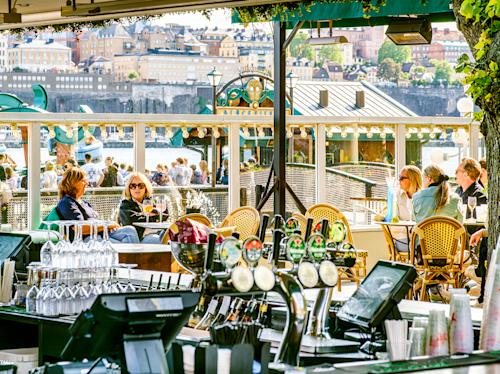 Outdoor bar and café with people sitting at tables and enjoying drinks, city buildings in the background, and sunlight brightening the scene.