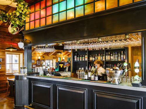 A bar with black wood panels, hanging wine glasses, bottles on shelves, and a bartender. Colorful stained glass panels decorate the top of the bar area.