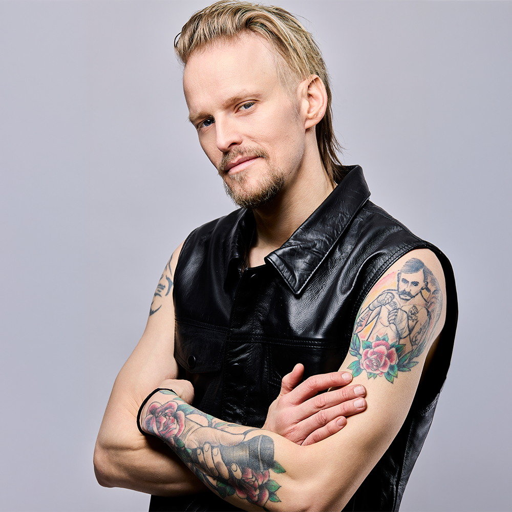 Man med blont hår och pipskägg, iklädd ärmlös svart skinnväst och med tatueringar på båda armarna, står med armarna i kors mot en enfärgad bakgrund.