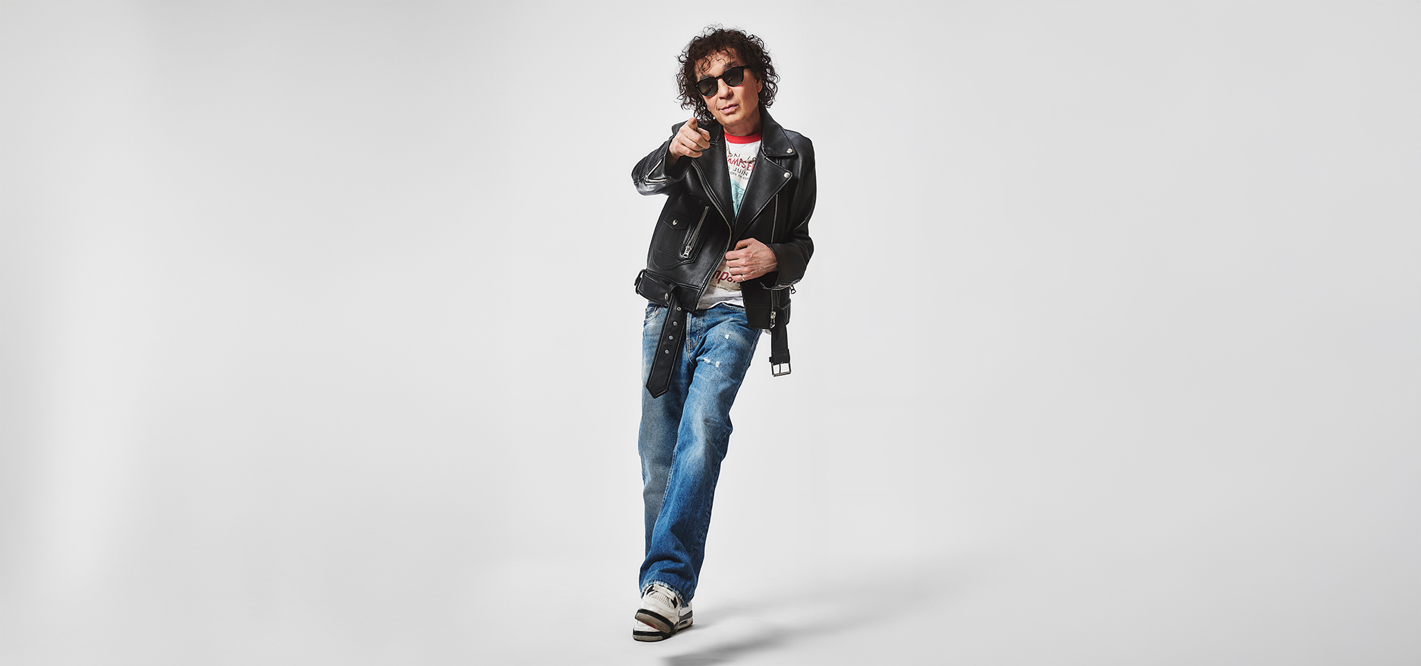En person som bär solglasögon, svart skinnjacka, jeans och sneakers pekar framåt och står mot en vit bakgrund.