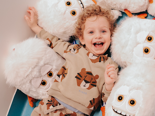 Ett litet barn med lockigt hår ler medan det ligger på golvet omgivet av fluffiga vita plyschleksaker med stora ögon och orangea fötter.