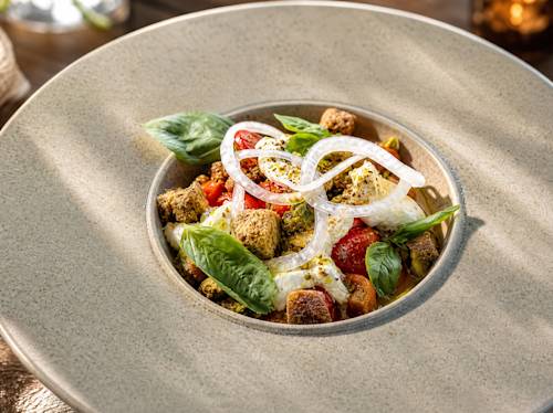 A bowl of burrata salad with cherry tomatoes, basil leaves, onion rings, and croutons, served on a textured beige plate.
