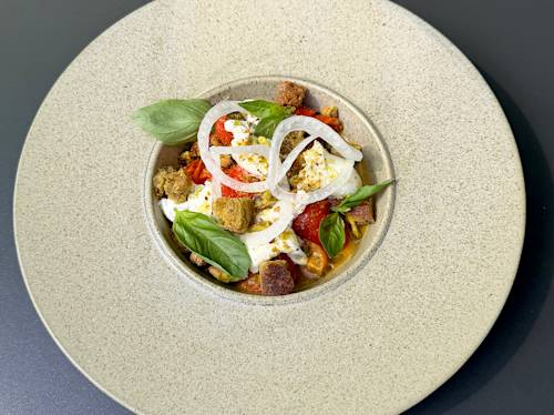 En upplagd sallad med körsbärstomater, burrataost, krutonger, färska basilikablad och lökringar, serverad i en beige skål på en mörk yta.