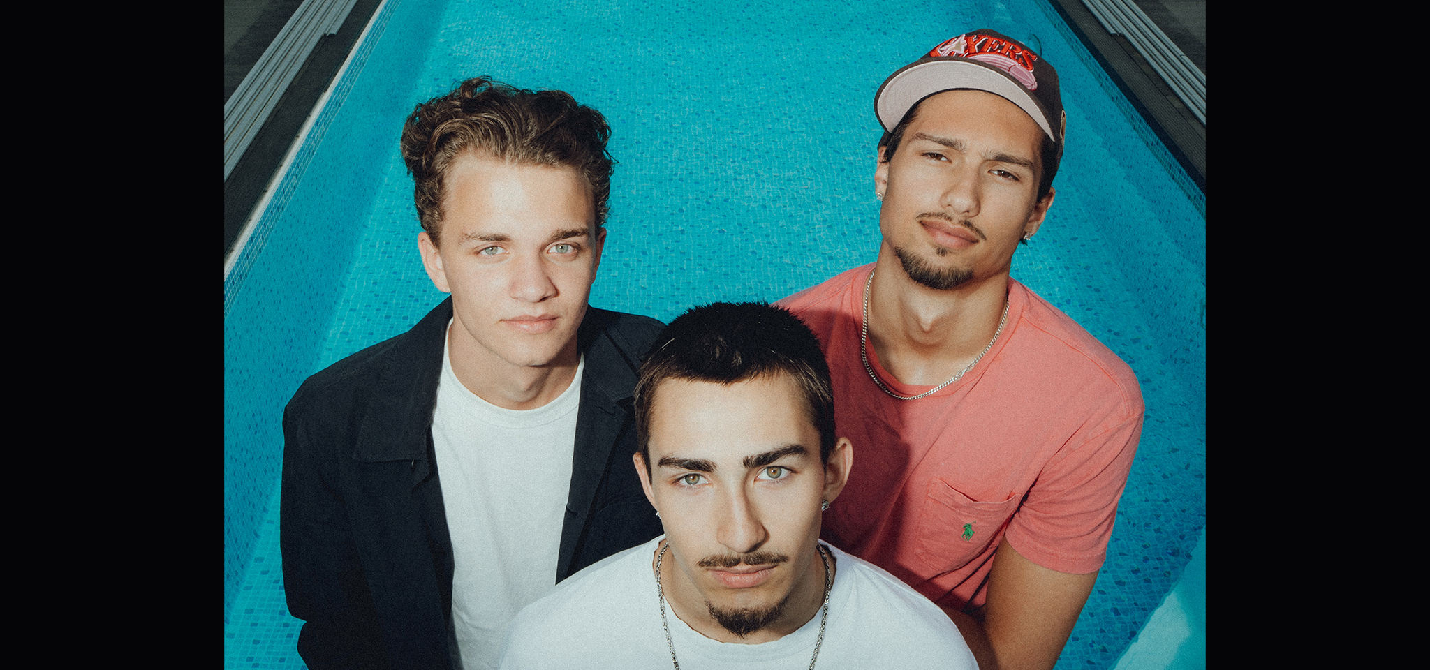 Three young men stand closely together at the edge of a blue swimming pool, looking up at the camera with neutral expressions.