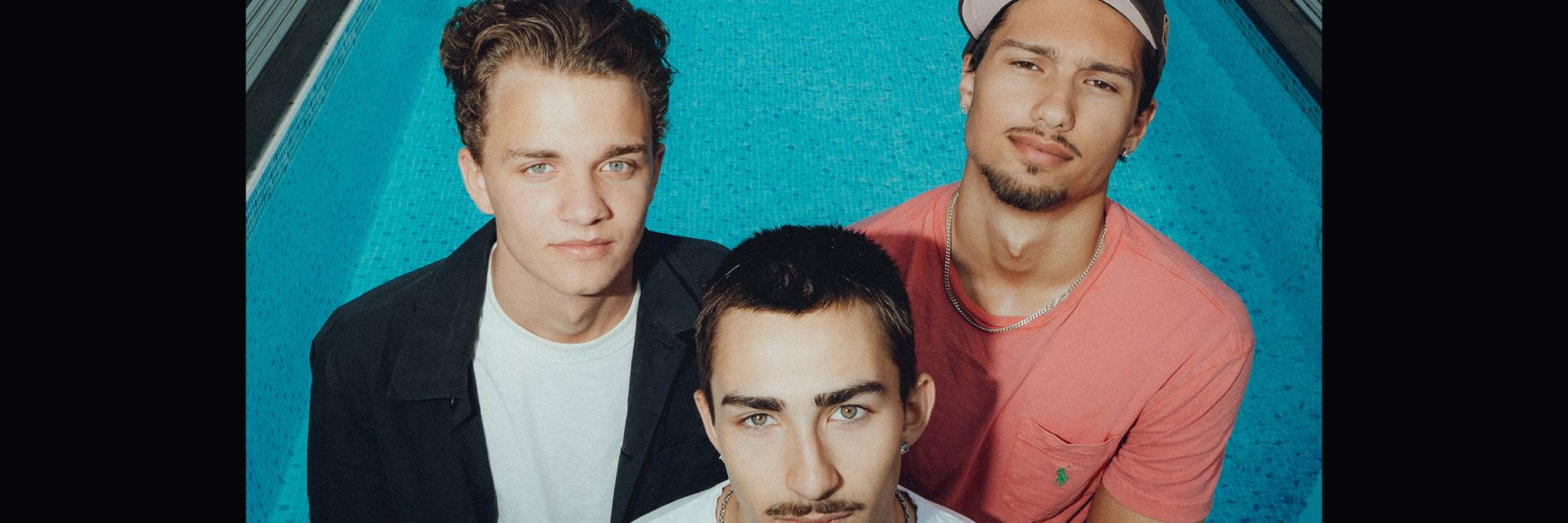 Three young men stand closely together at the edge of a blue swimming pool, looking up at the camera with neutral expressions.