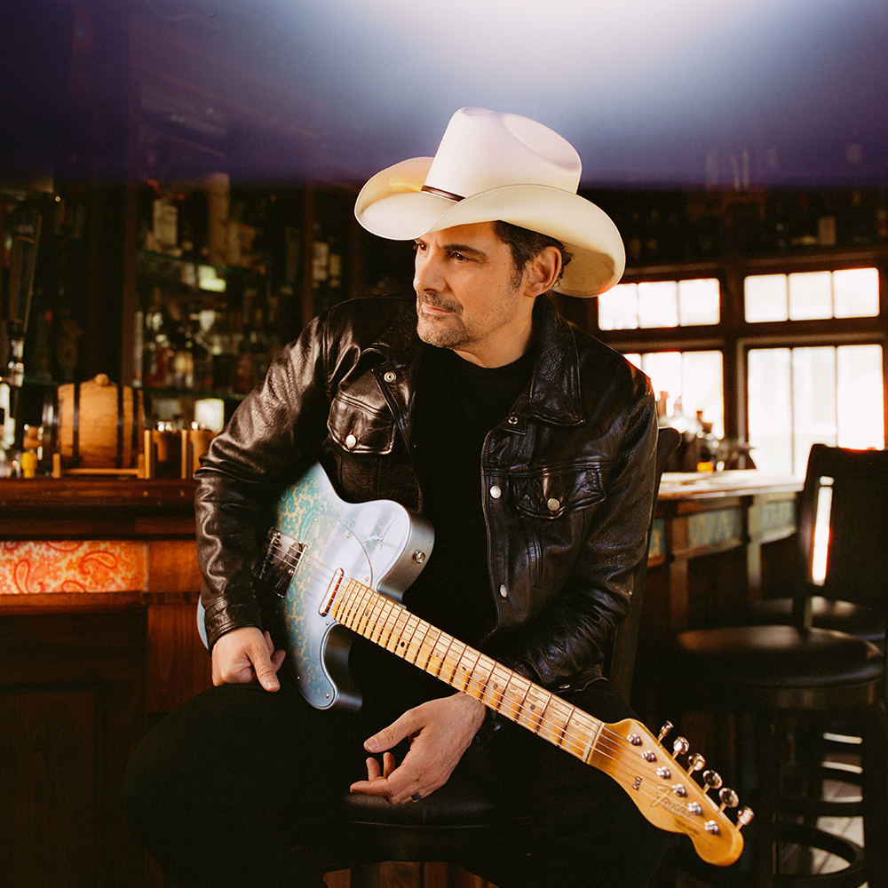 A man in a black leather jacket and white cowboy hat sits on a barstool holding an electric guitar in a dimly lit bar.