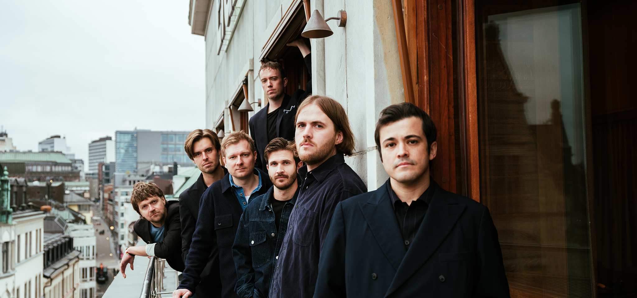 Sju män står på en balkong med utsikt över en stadsbild, vända mot kameran med allvarliga ansiktsuttryck. Stadens byggnader syns i bakgrunden under en mulen himmel.