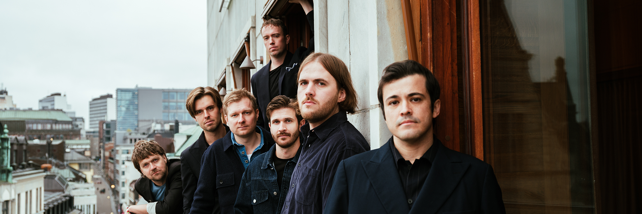Sju män står på en balkong med utsikt över en stadsbild, vända mot kameran med allvarliga ansiktsuttryck. Stadens byggnader syns i bakgrunden under en mulen himmel.