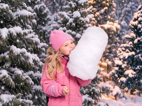 En liten flicka i en rosa vinterrock och mössa låtsas äta en stor snöboll framför snötäckta barrträd.