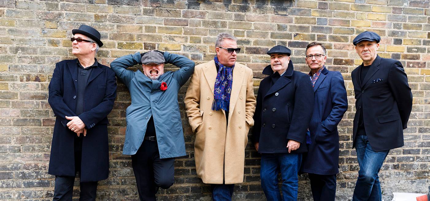 Six men in coats and hats stand against a brick wall, some with hands in pockets and one with hands on head, posing casually.