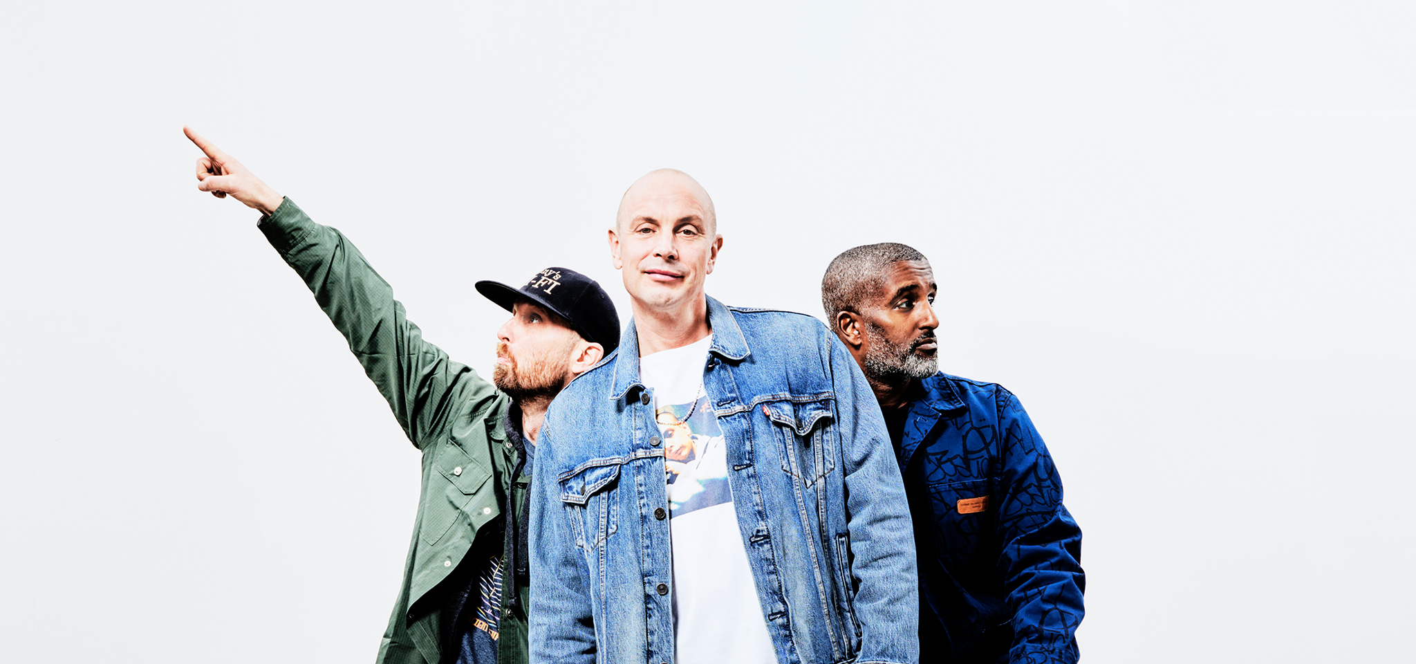 Tre män poserar mot en vit bakgrund; en pekar åt vänster, den mellersta står vänd framåt och den tredje tittar åt höger.