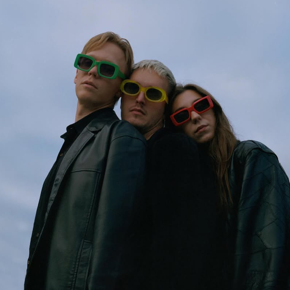 Three people wearing dark jackets and colorful sunglasses stand closely together against a cloudy sky.