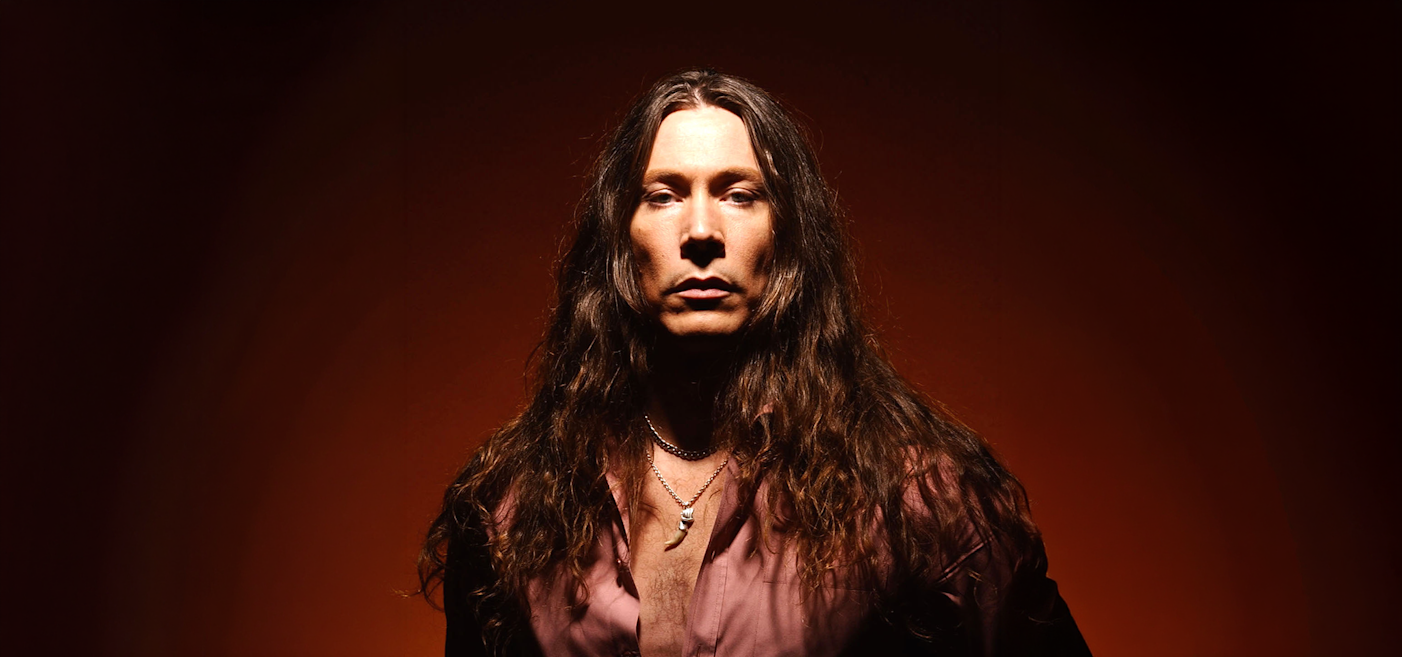 En person med långt brunt hår, iklädd en nedknäppt skjorta och flera halsband, står mot en mörkorange bakgrund.