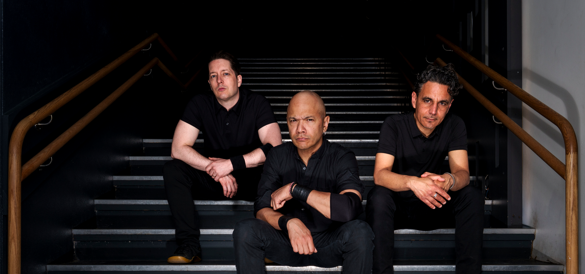 Three men dressed in black sit on stairs inside a building, each with a serious expression, facing the camera.