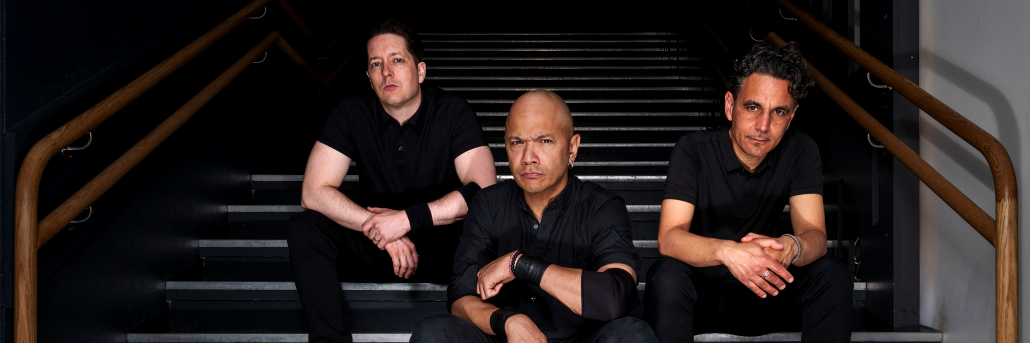 Three men dressed in black sit on stairs inside a building, each with a serious expression, facing the camera.