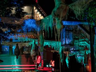 Nattscen utomhus med byggnader dekorerade med trasigt tyg och kuslig belysning som skapar en spöklik atmosfär med Halloween-tema. Neonskyltar syns i bakgrunden.