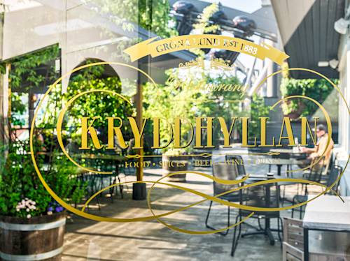 Gold-lettered window of Restaurang Kryddhyllan at Gröna Lund, reflecting outdoor seating, greenery, and a few people sitting at tables in the sunny patio area.