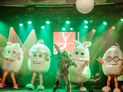 Four people in large white fuzzy mascot costumes are on stage with a performer in a green outfit, under green lights, surrounded by Halloween decorations.