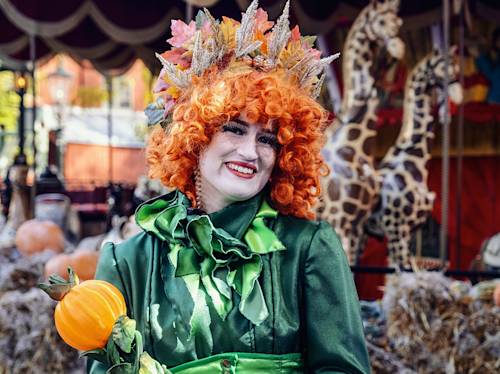 Person in a green costume with an orange curly wig and autumn leaf crown holds a small pumpkin, standing in front of hay bales and giraffe figures at a festive outdoor event.
