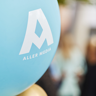 A blue balloon with the white Aller Media logo and text. The background is blurred with indistinct people and shapes.