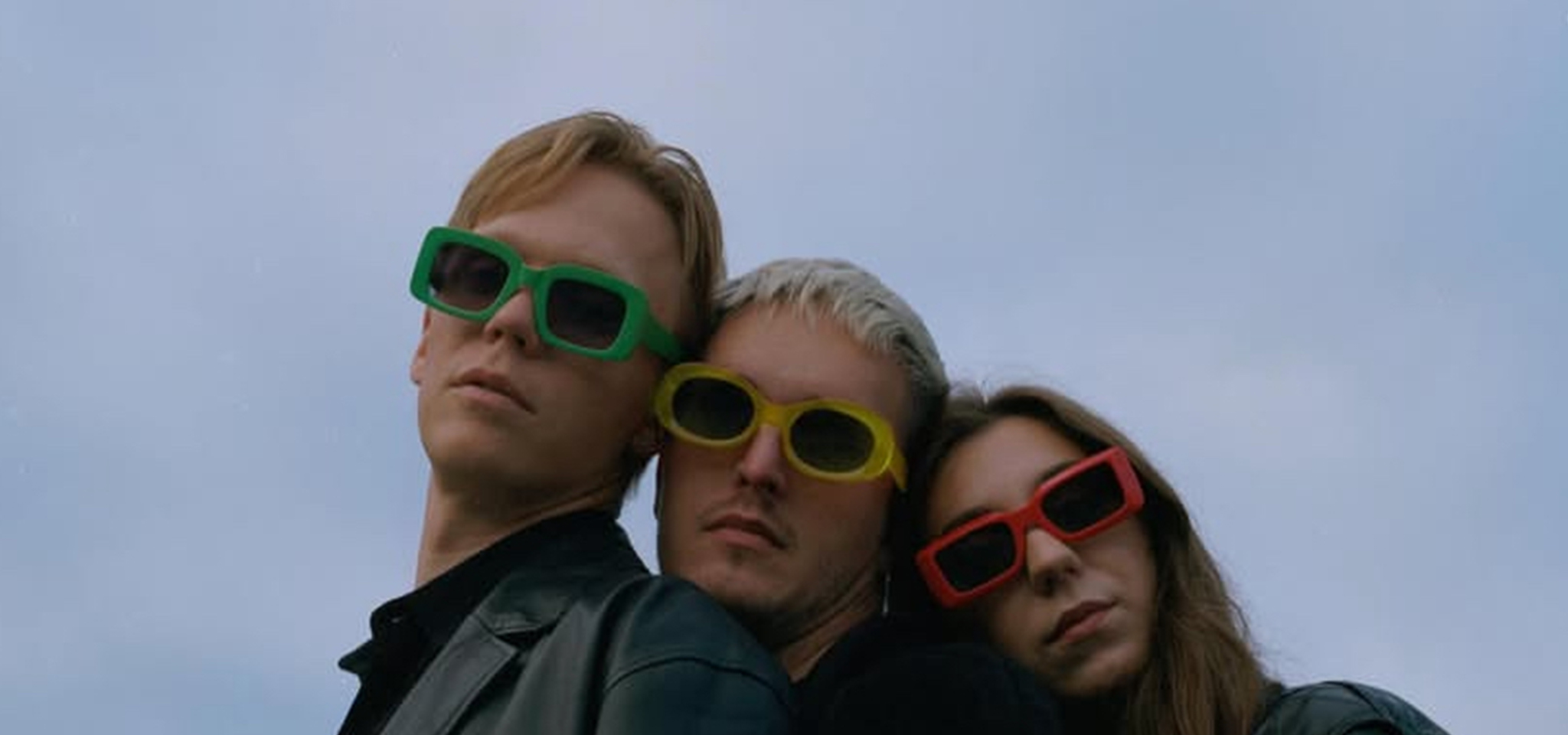 Three people wearing colorful sunglasses and black jackets stand close together against a cloudy sky.
