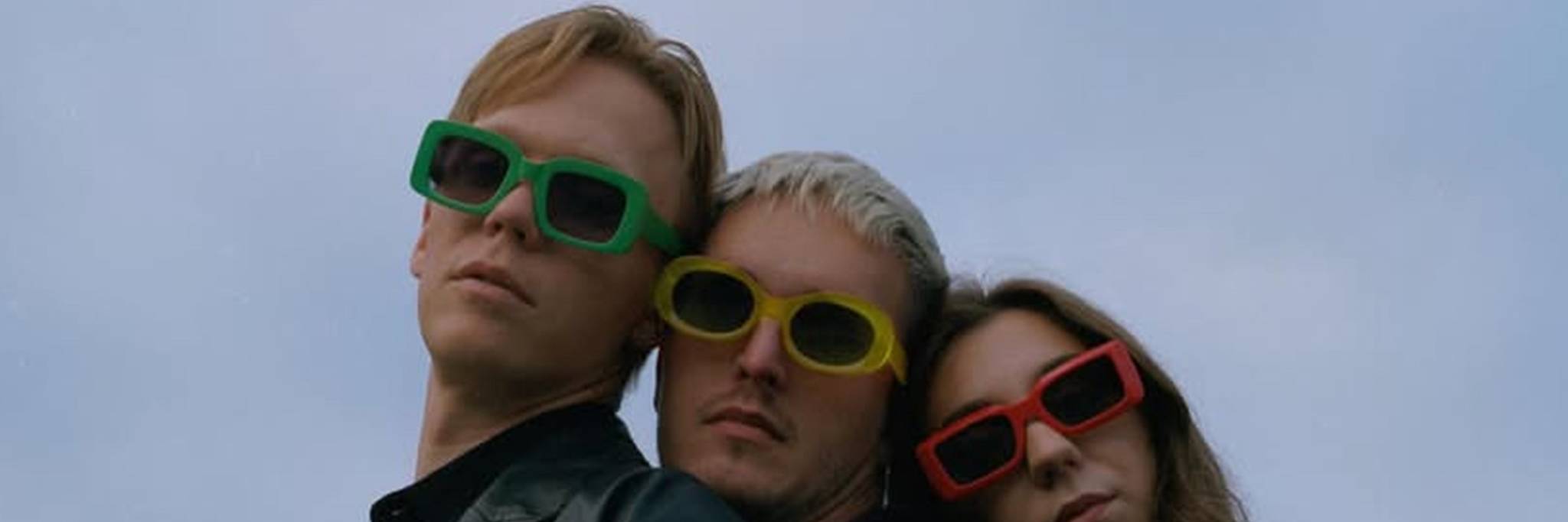 Three people wearing colorful sunglasses and black jackets stand close together against a cloudy sky.