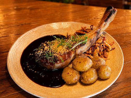 En tallrik med en tomahawk-fläskkotlett toppad med örter och ströbröd, serverad med ugnsbakade potatisar, mörk sås och en sidorätt av tunt skurna stekta grönsaker.