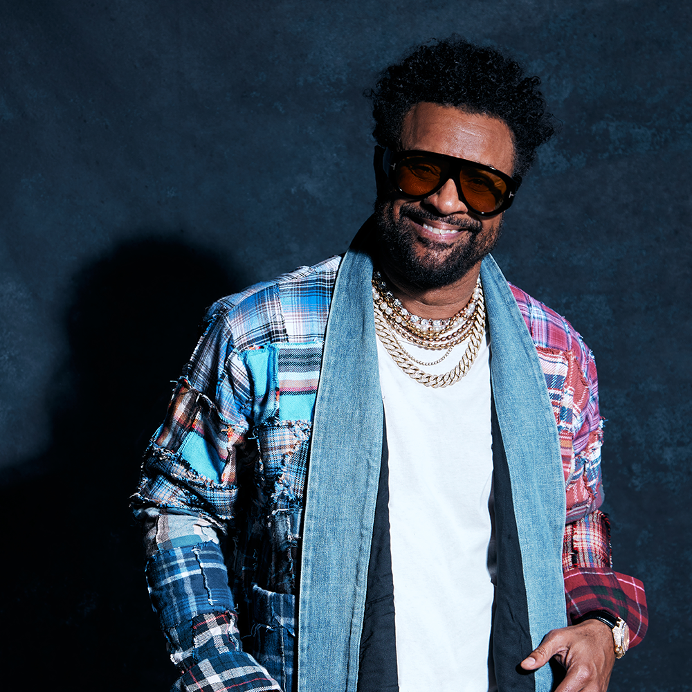 En man med solglasögon, halsband i flera lager, vit t-shirt, patchworkjacka och jeanshalsduk ler mot en mörk bakgrund med textur.