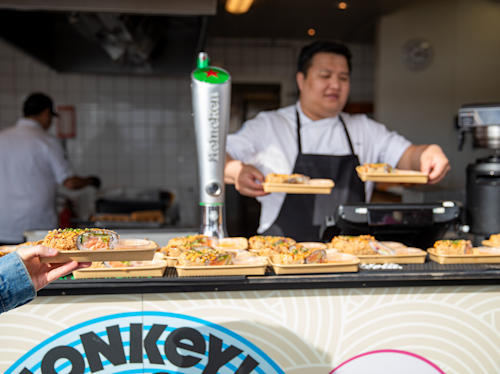 A chef hands plates of sushi to customers at a food stall, with a Heineken beer tap visible in the background.